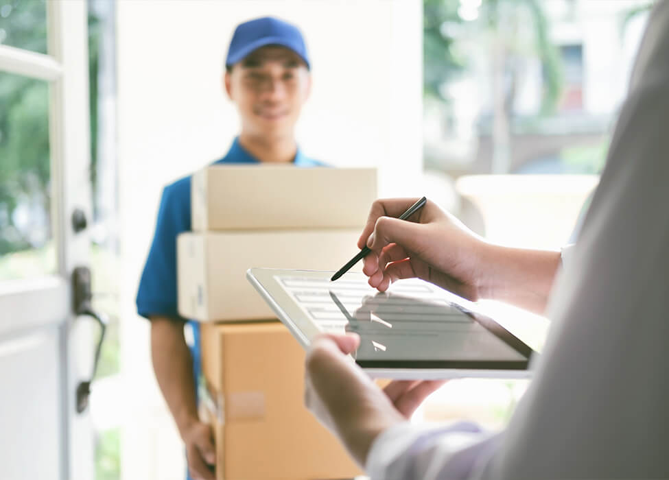 A man in a courier uniform holds packages at another person's door while the person signs a tablet