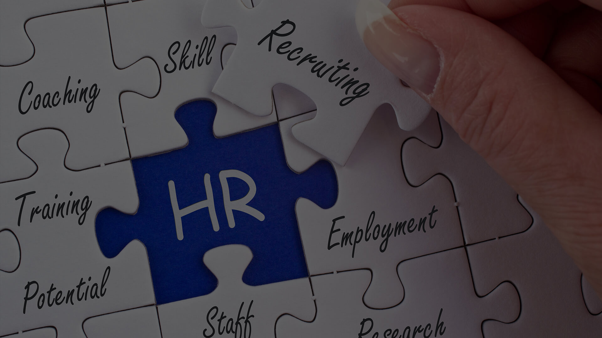 Harnessing Human Resources Services for Business Growth The image is a close-up photograph of a person's hand completing a jigsaw puzzle. The puzzle pieces are white with printed words that are related to human resources (HR) functions and attributes. The visible words include "Coaching," "Skill," "Recruiting," "Training," "Potential," "HR," "Employment," "Staff," and "Research." The piece being placed by the hand is the final piece and carries the acronym "HR," which connects to the blue piece already in place with the matching protruding part. The overall the