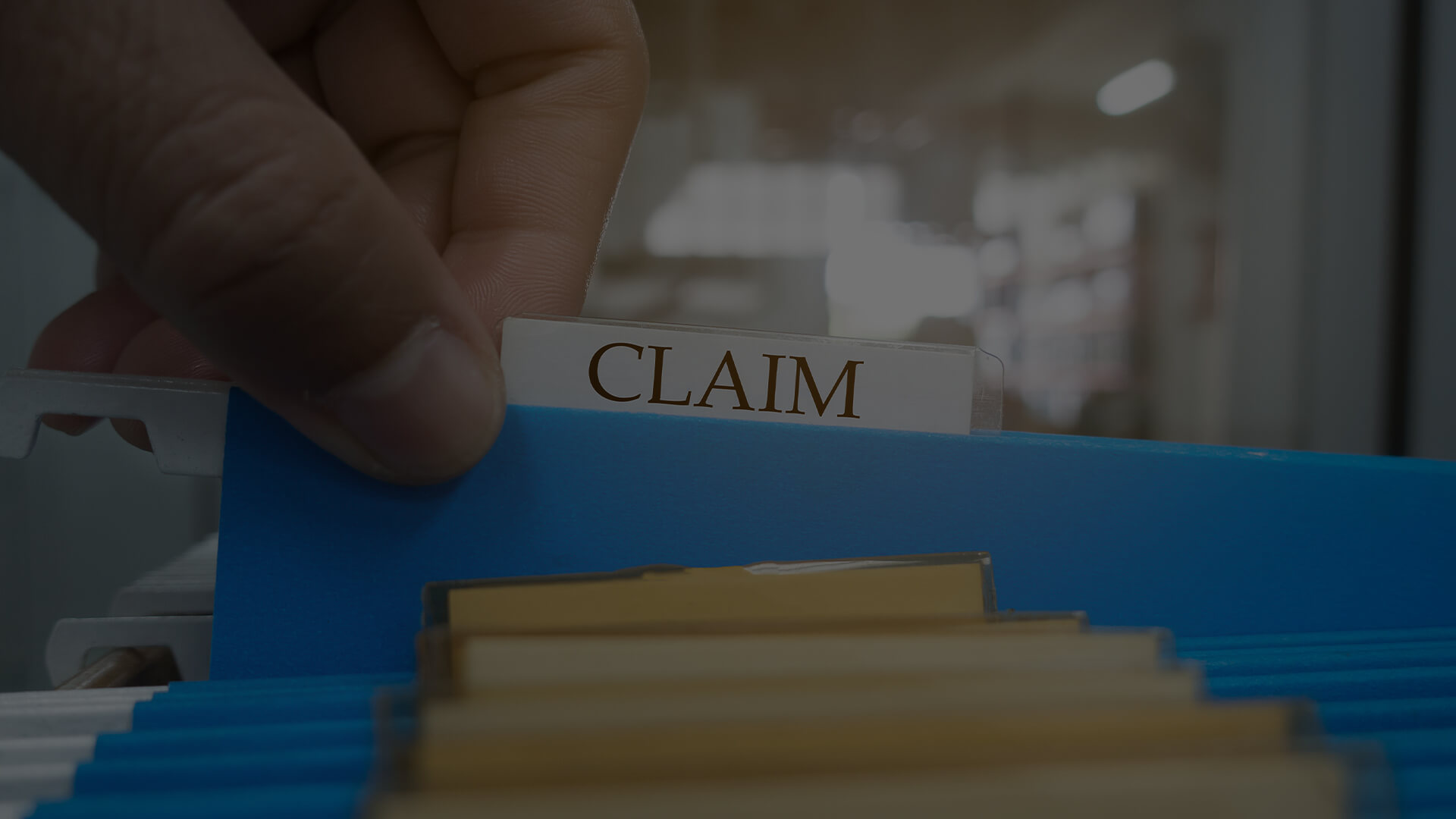 A close-up of a hand pulling out a blue file folder labeled "CLAIM" from a filing cabinet, representing the medical claims process. The background shows a blurred office setting with rows of similar files