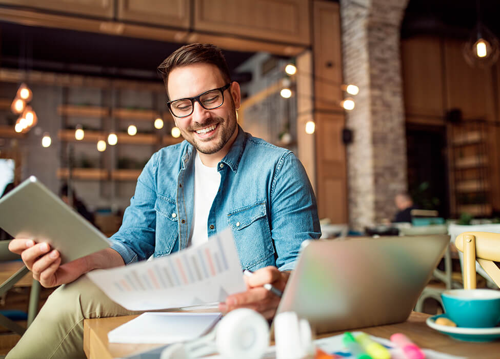 In a coffeeshop, a man smiles as he looks at papers, has his laptop in front of him and has papers scattered on the table