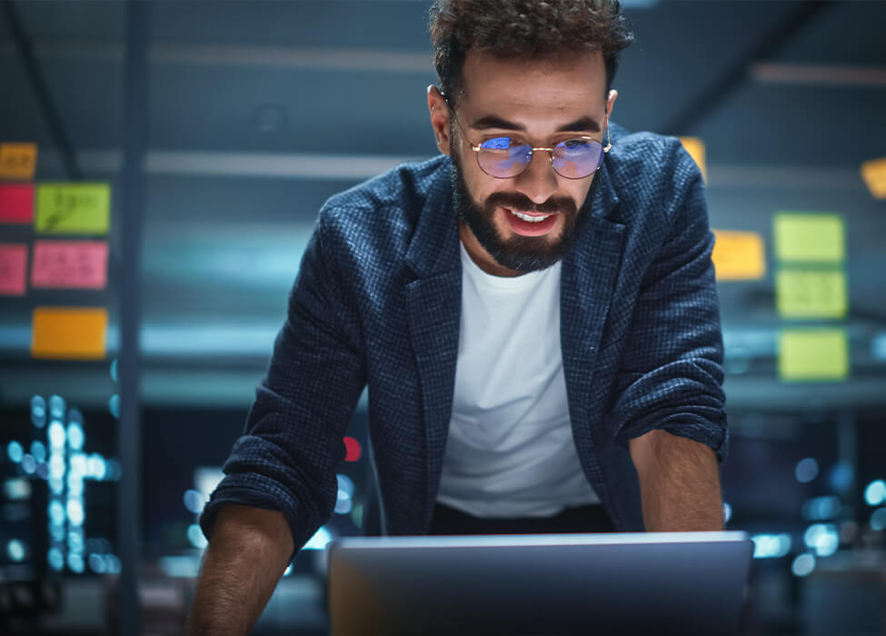 Man stands in a computer room and smiles down at his laptop