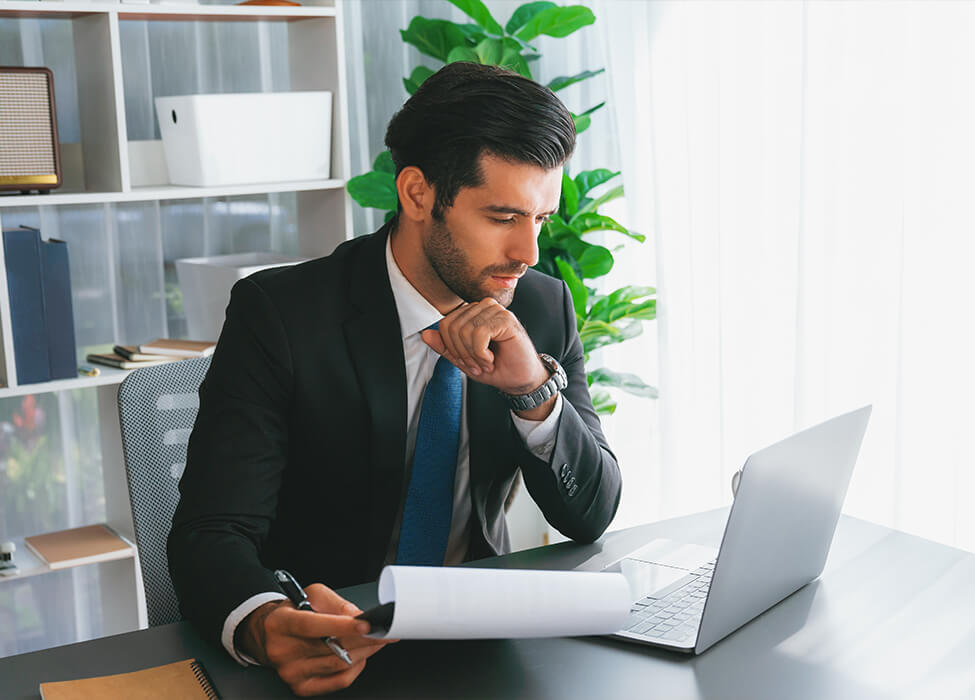 Man in suit looks at his laptop while holding papers in one hand