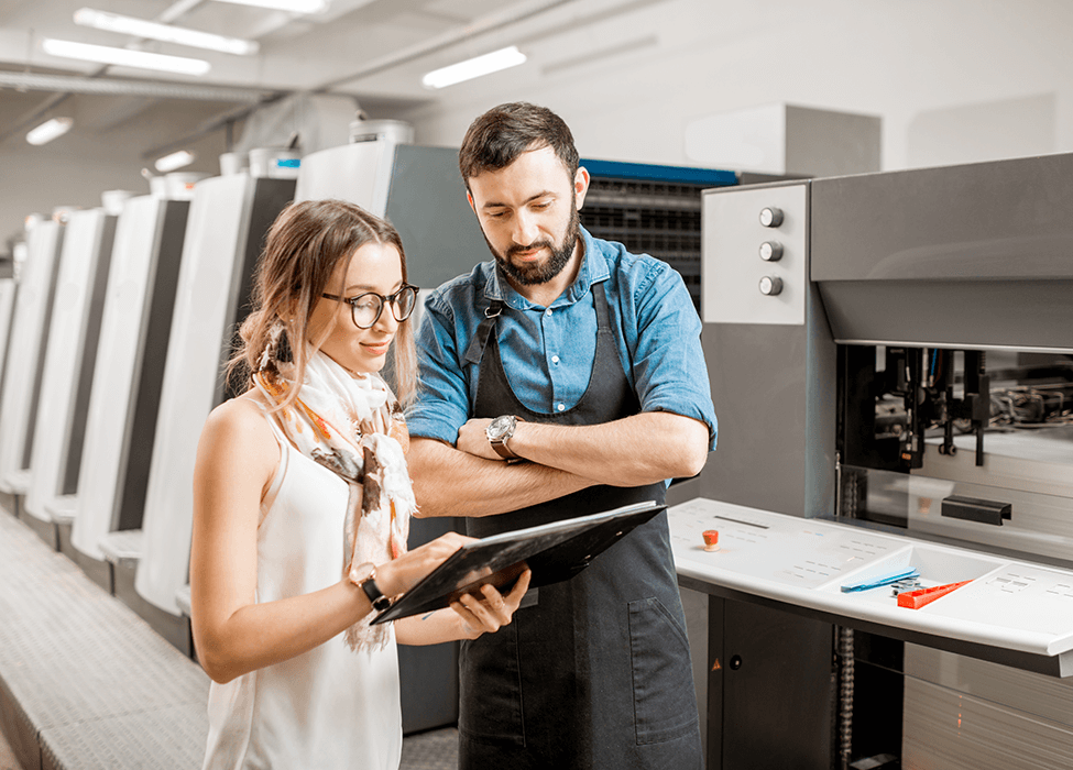 A male and female professional discussing over a digital tablet in a modern printing facility, indicating the consultation process in managed print services.