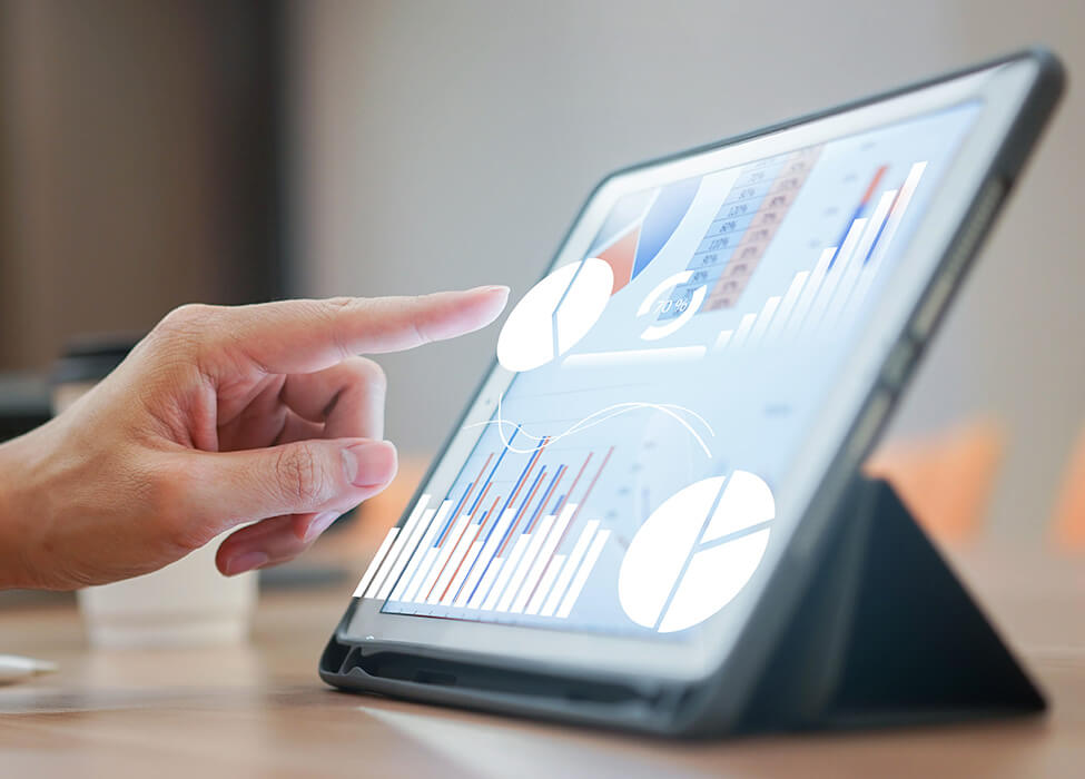 Close-up of a hand pointing to a colorful pie chart on a tablet screen, which is resting on a stand on a wooden desk. The tablet displays various financial charts and graphs, indicating a user engaged in digital analysis or review of financial data and metrics