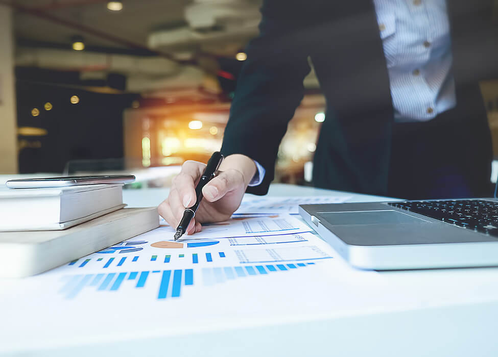 A business professional is analyzing financial charts and data on paper documents laid out on a table. Next to the papers is a laptop and a smartphone, suggesting a hybrid approach to data analysis combining traditional and digital methods in a business setting.