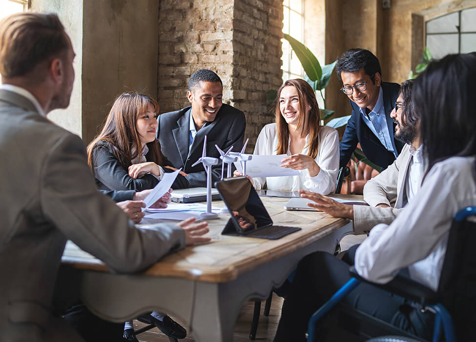 A diverse group of business professionals engaging in a meeting, discussing sustainable business practices with models of wind turbines on the table.